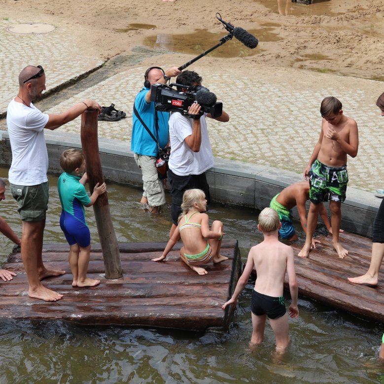 Kinder spielen auf dem Wasserspielplatz auf der Schlossinsel und werden von einem Kamerateam gefilmt.
